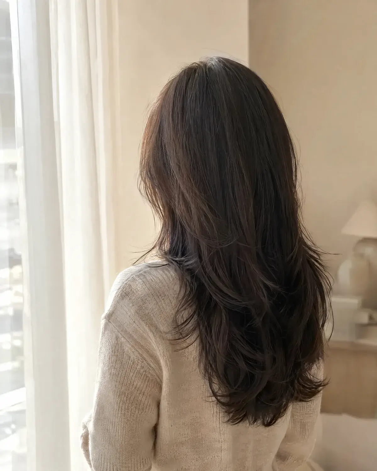창가에 선 여성의 뒷모습, 어깨 아래로 층이 흐르는 긴 레이어드 머리