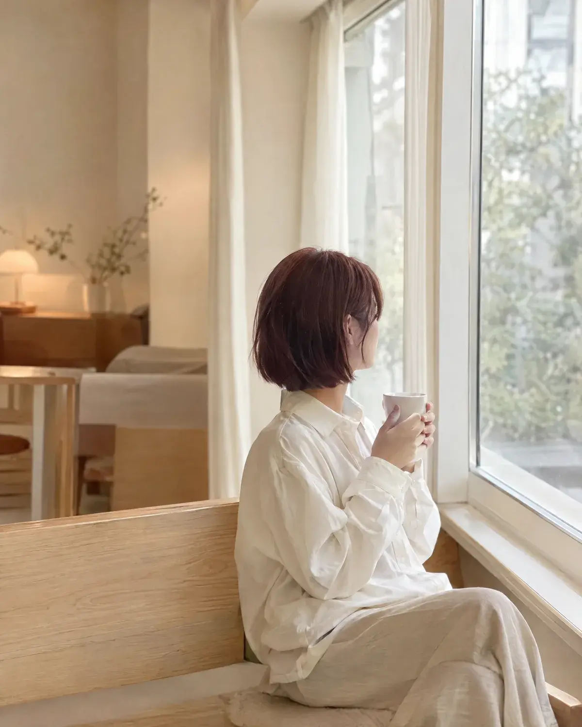 차분한 버건디 갈색 단발에 아이보리 셔츠를 입고 흰 컵을 든 채 창밖을 보는 여성의 옆모습